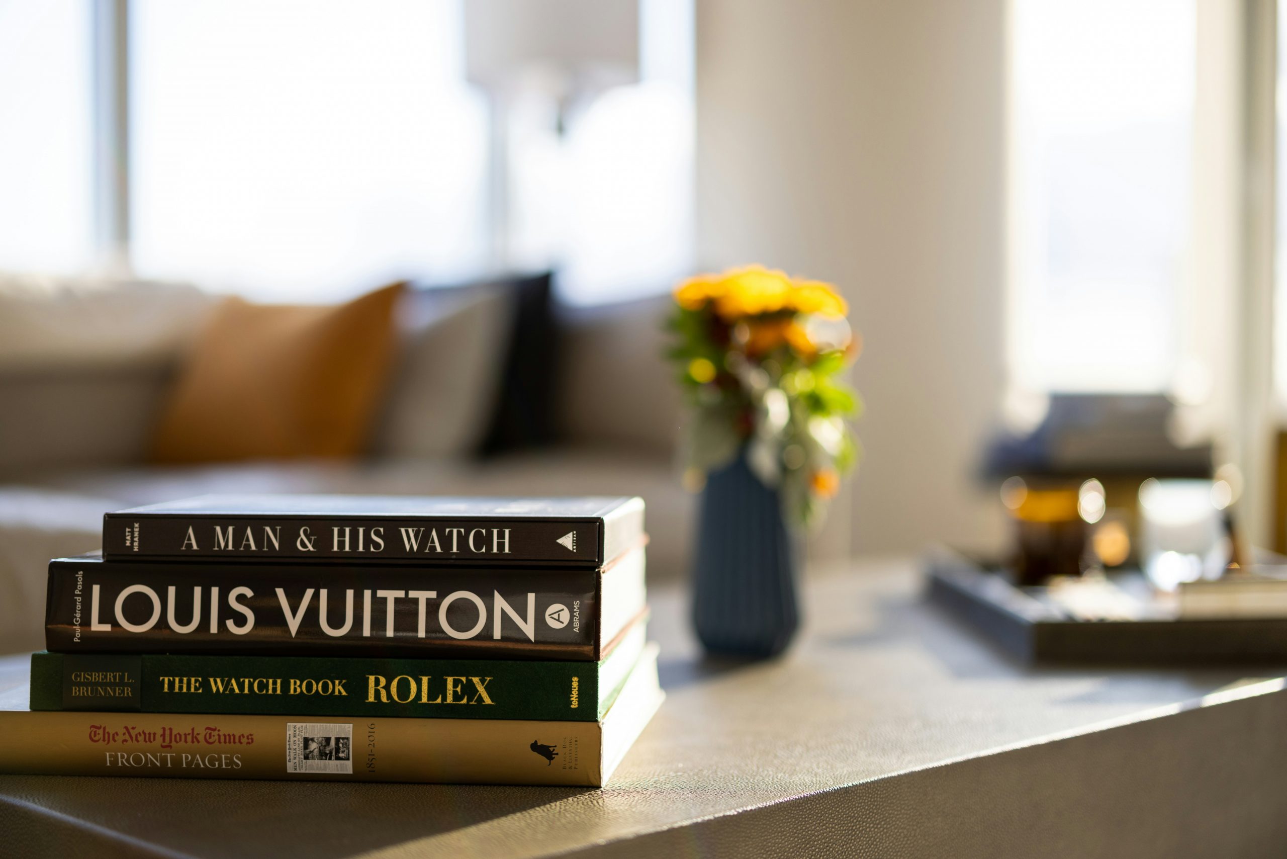 A stack of luxury coffee table books including Louis Vuitton and Rolex, placed on a modern table with blurred background decor.