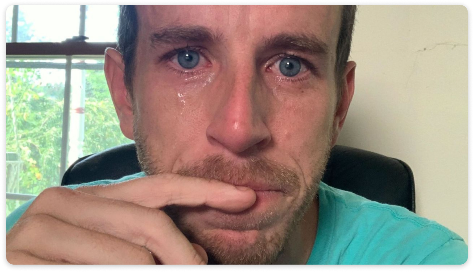 Close-up photo of a man with teary eyes sitting indoors, resting his hand near his mouth.
