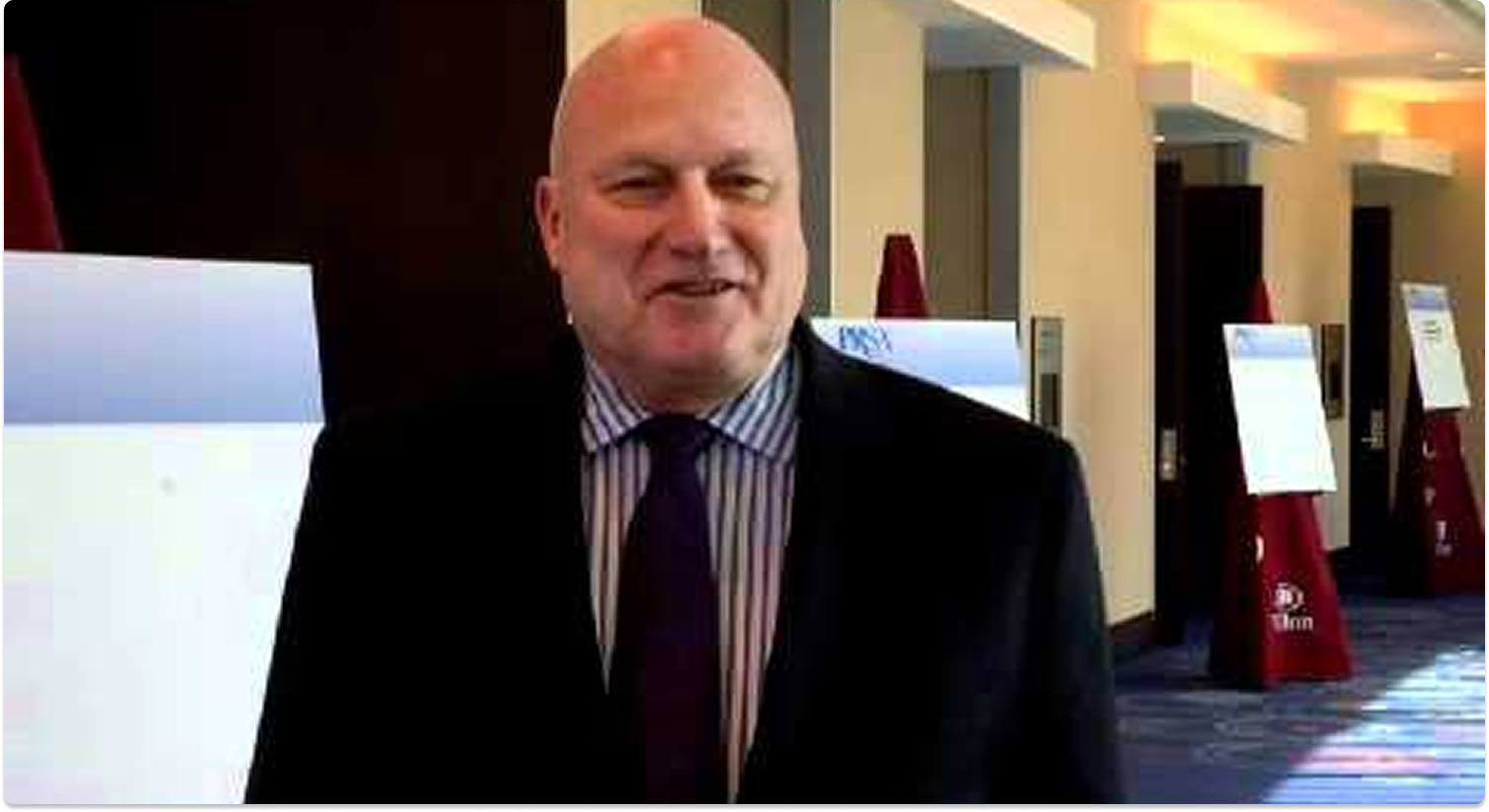 Man in a suit speaking at a conference venue with presentation boards in the background.