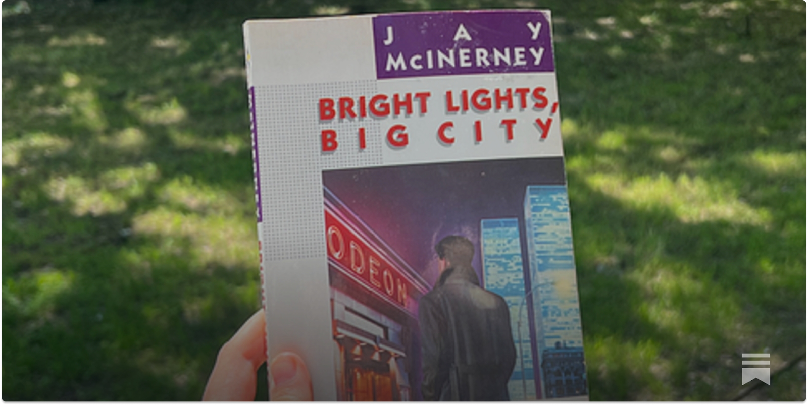Hand holding the book “Bright Lights, Big City” by Jay McInerney outdoors in a park.